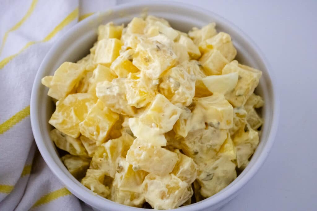 Southern Potato Salad Recipe (My Mom's Famous Recipe) 3 Overhead view of Southern potato salad in a white serving bowl, white and yellow checkered kitchen towel on a white countertop.