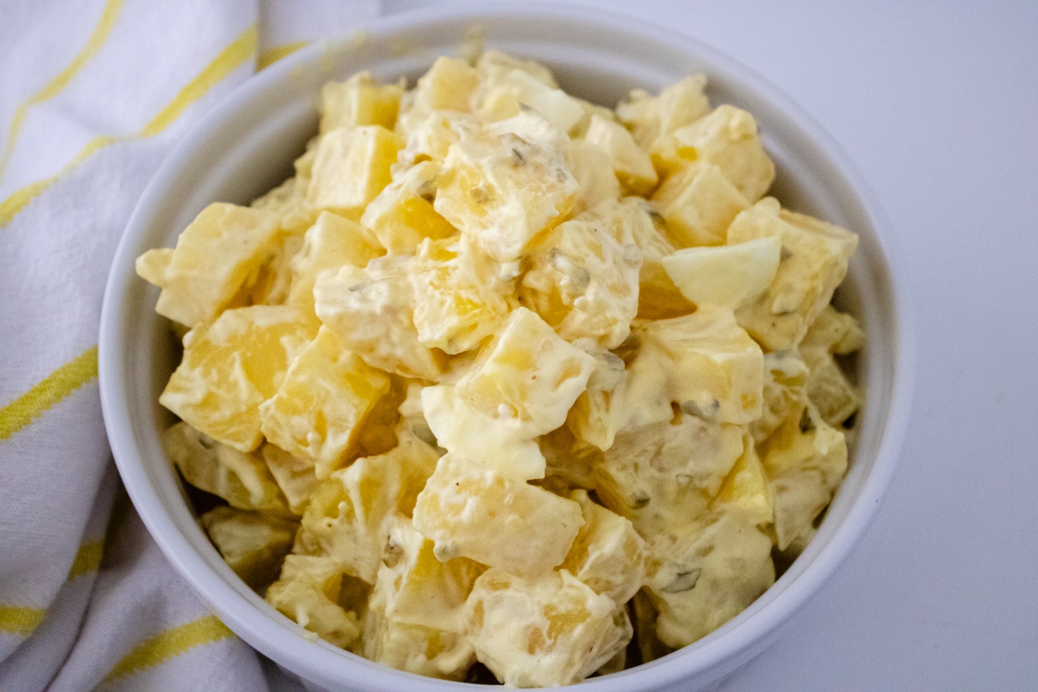 A white ceramic dish filled with creamy potato salad, featuring chunks of potatoes coated in a yellow dressing. In the background, there's a white and yellow striped cloth. The overall setting is minimalistic with a light-colored backdrop.