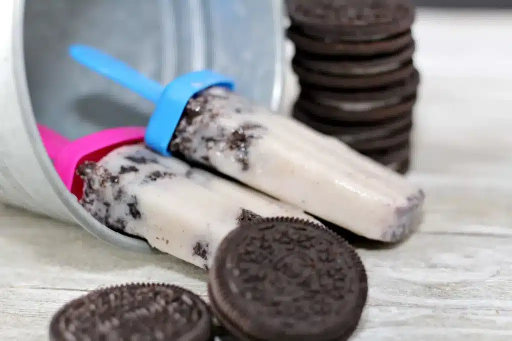 Two Cookies and Cream Popsicles with pink and blue handles at its bases. The popsicles are placed in a small tin bucket that has toppled over.