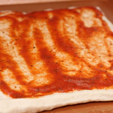 A square pizza dough slathered with tomato sauce.