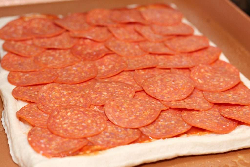 Focus shot on a square pizza dough for the Spicy Stuffed Pizza Bread topped with tomato sauce and thinly sliced pepperoni.