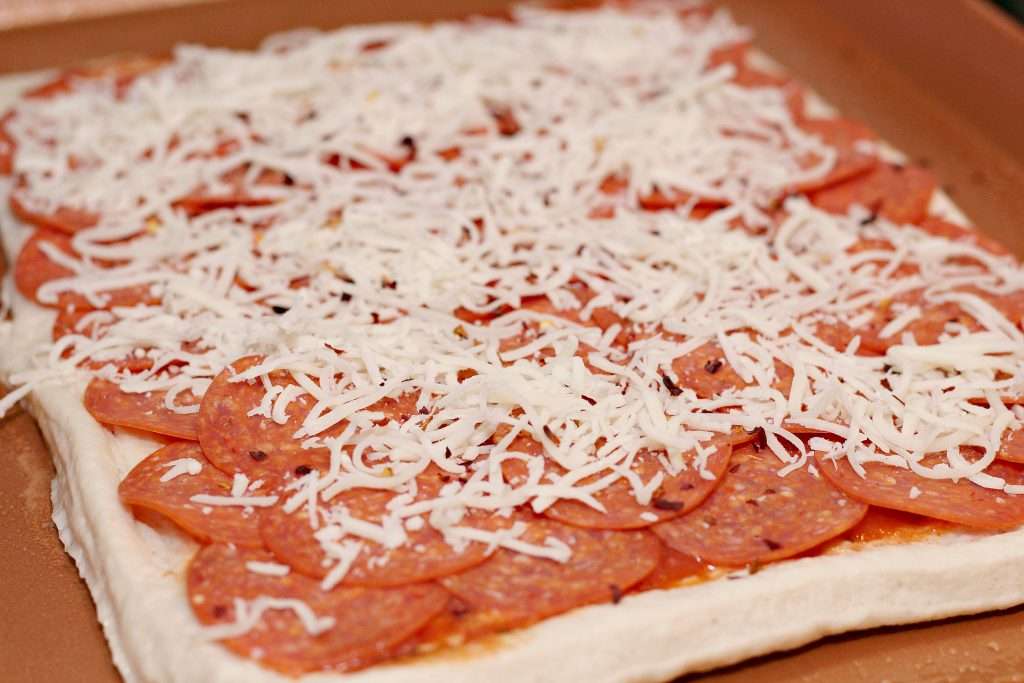 Focus shot on a square pizza dough for the Spicy Stuffed Pizza Bread topped with tomato sauce and thinly sliced pepperoni. Shredded mozzarella cheese has been added as well.