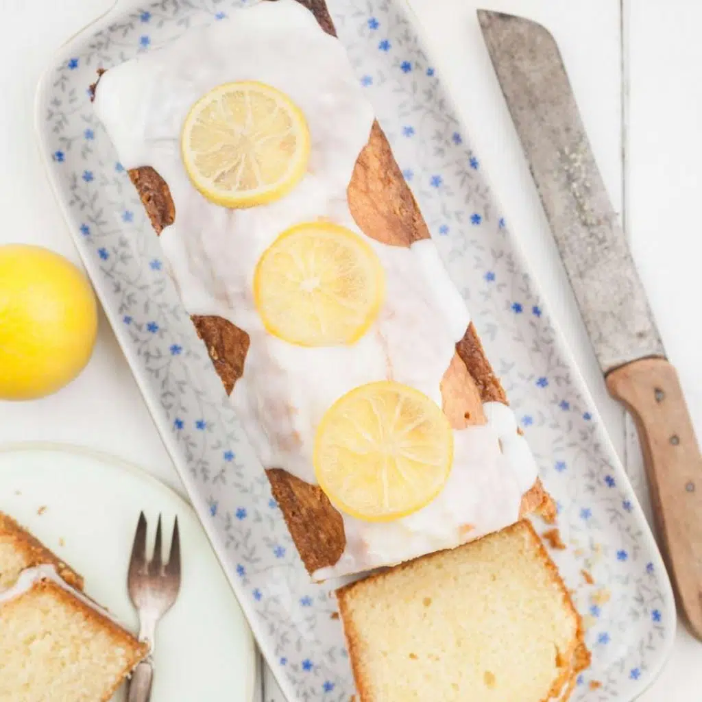 Flat lay shot of the finished Southern Lemon Pound Cake loaf, topped with lemon sugar glaze and lemon slices on it. The loaf is placed on a rectangular platter with blue floral designs on it. The platter is then placed on a white wooden surface, surrounded by whole lemons and a bread knife.