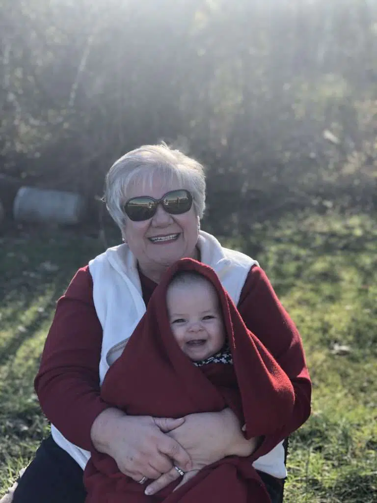 A woman wearing a red sweater and a white vest over it, and sunglasses, happily carrying a baby with a red blanket on her lap. She and the baby are sitting outdoors. 