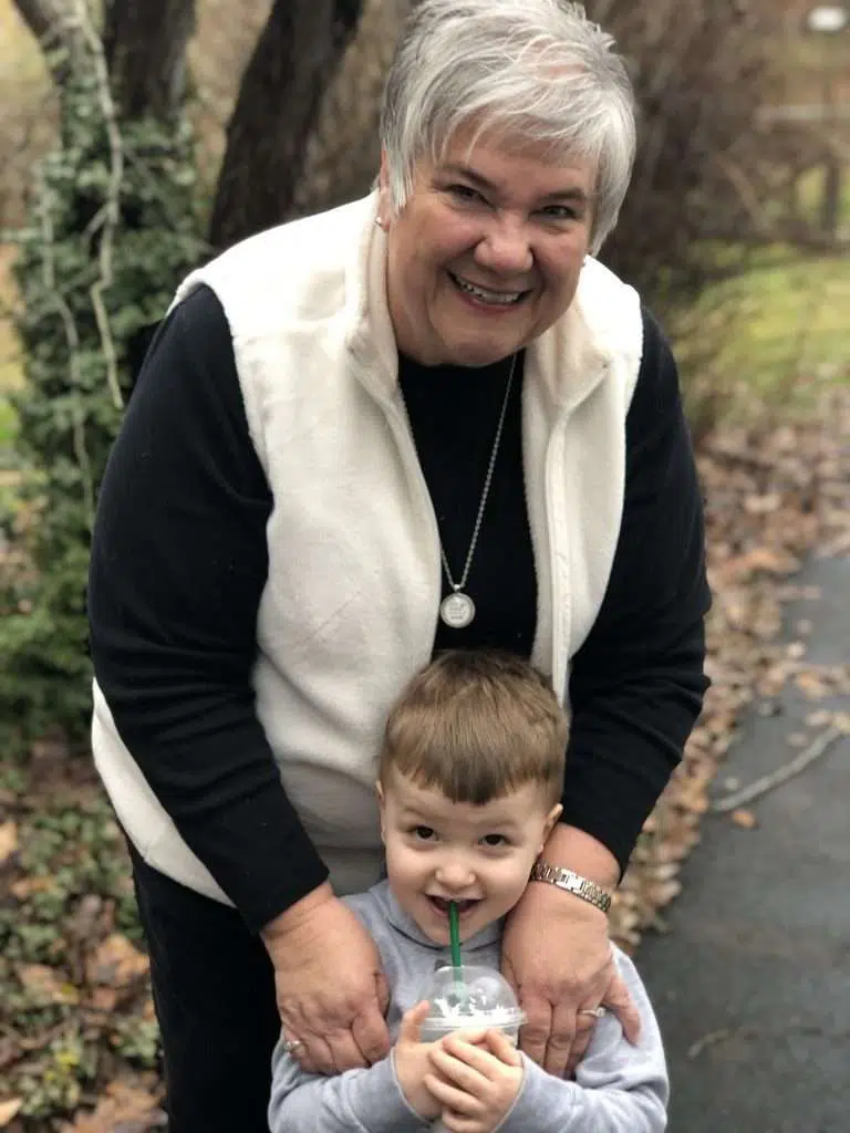 A woman wearing a black sweater with a white vest over it, while fondly holding her grandson by the shoulders as they smile for a picture.