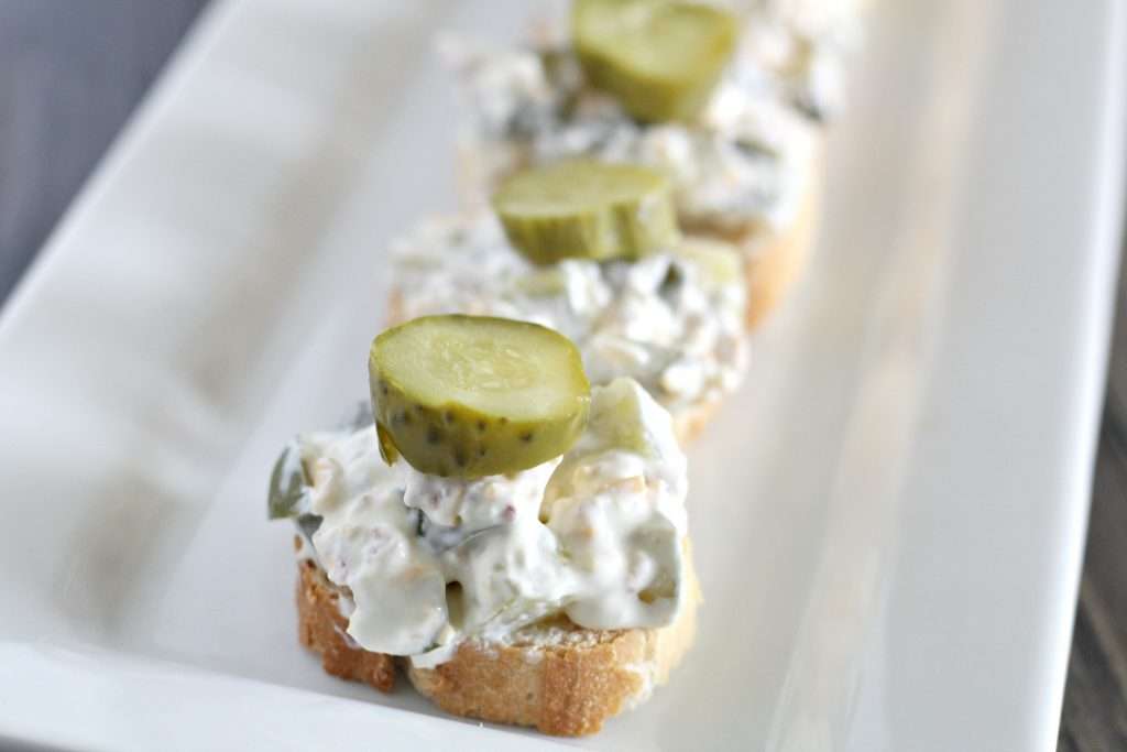 dill pickle dip on french bread on white platter topped with dill pickles