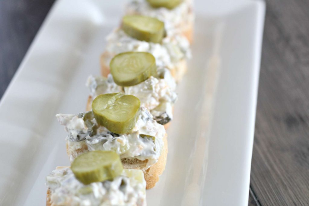 Delicious and Easy Dill Pickle Dip 2 dill pickle dip on a piece of bread on a white platter topped with a sliced pickle