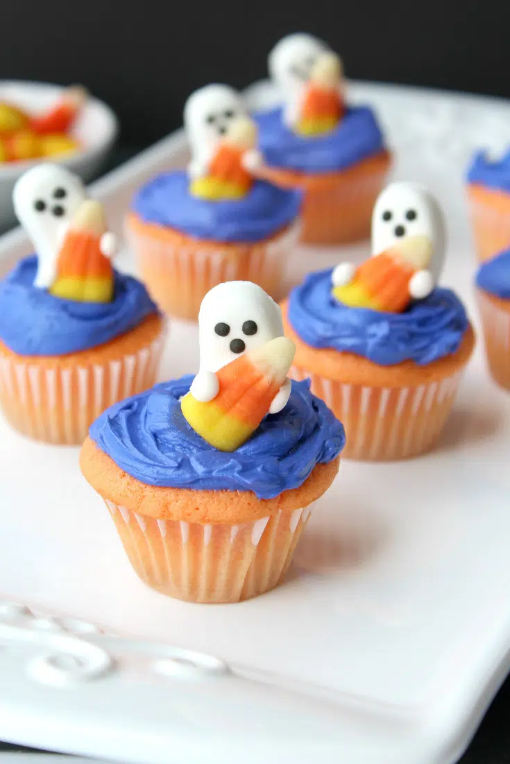 Ghost shaped candy on top of blue frosted cupcakes on a serving white plate.