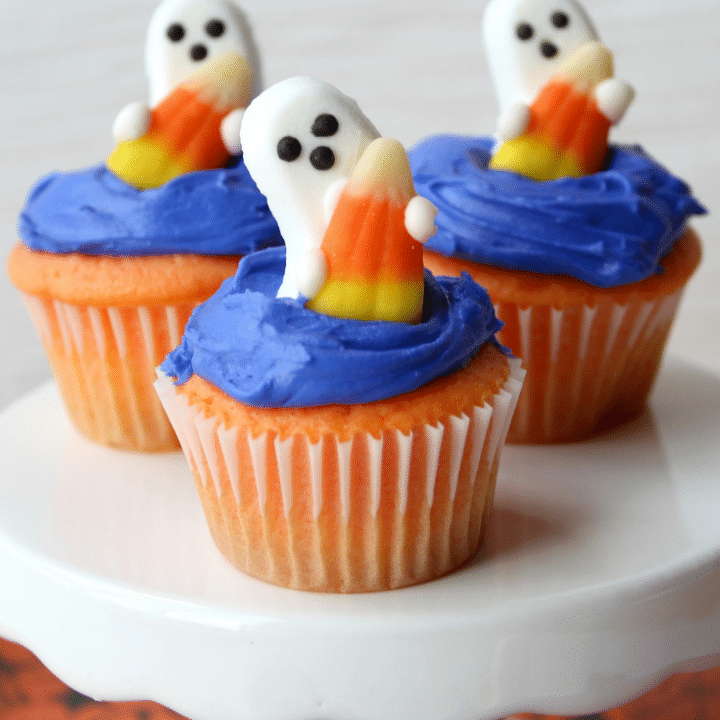 ghost shaped candy on top of blue frosted cupcakes on a white cake stand.