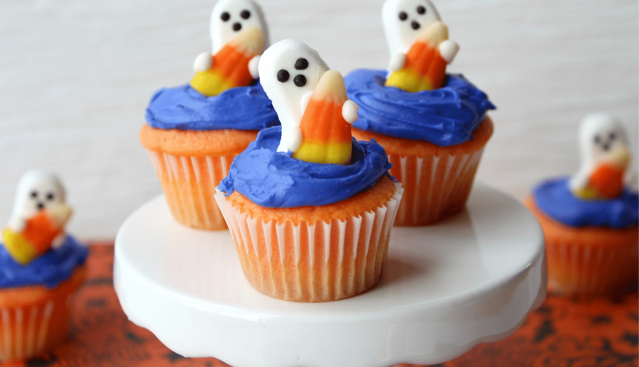 ghost shaped candy on top of blue frosted cupcakes on a white cake stand.