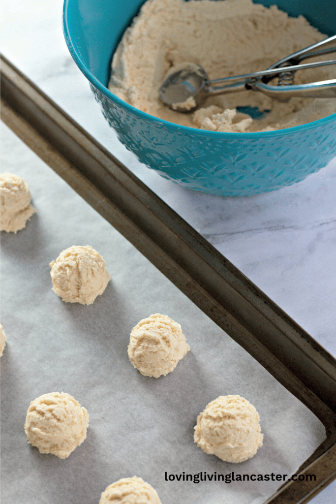Amish Sugar Cookies 14 Amish sugar cookie dough balls on a baking sheet ready to be baked, mixing bowl with dough and a cookie scoop next to the baking sheet.