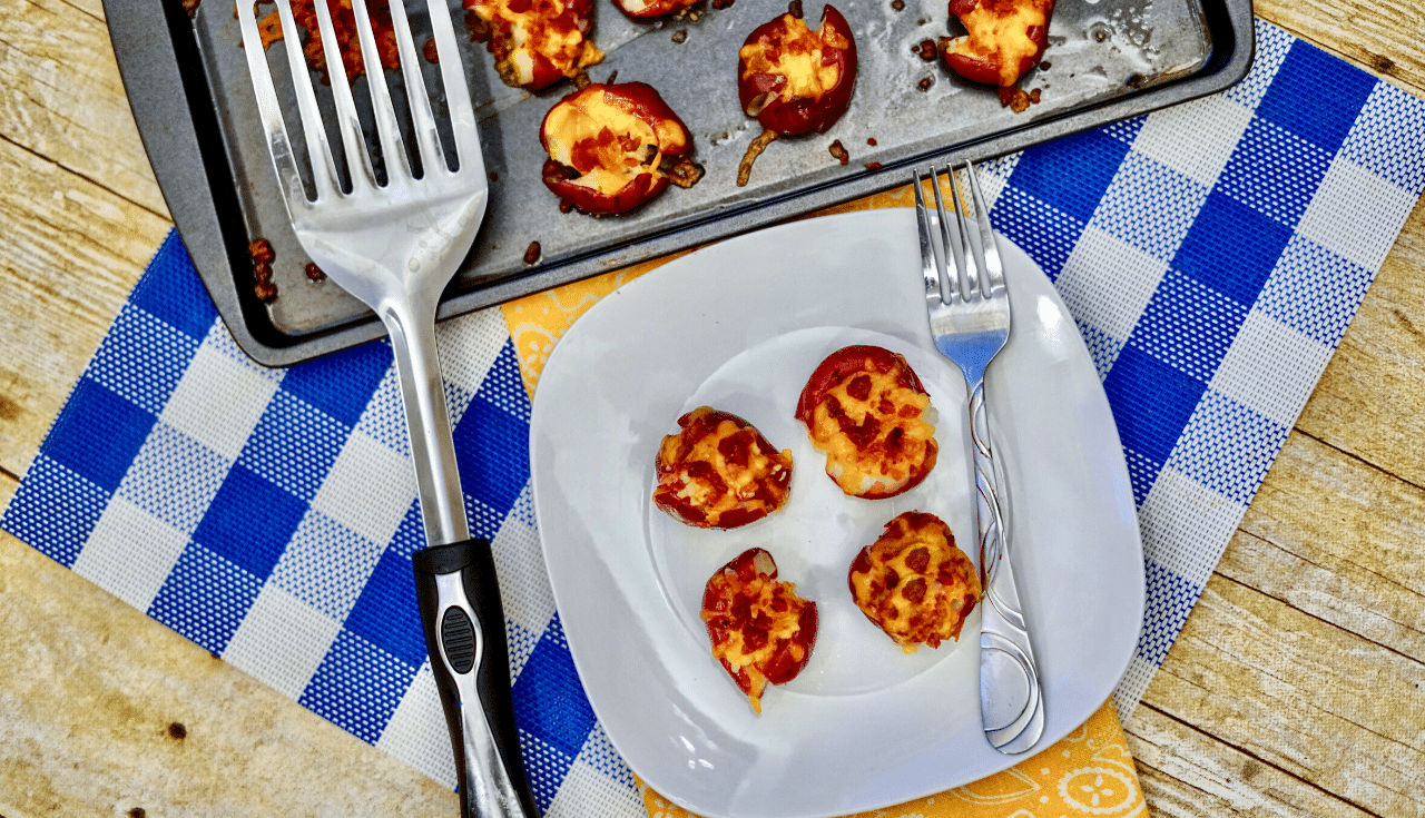 A white plate with four mini pizza bites, resembling loaded smashed potatoes, next to a fork, beside a baking tray with more pizza bites and a spatula, on a blue checkered cloth.