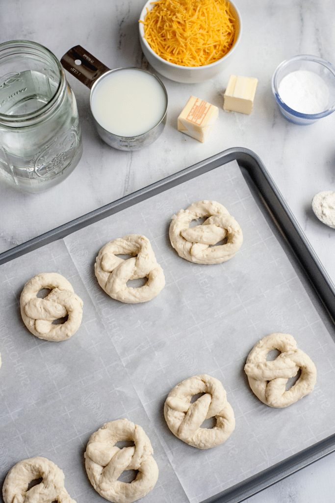 Soft Pretzels with Biscuit Dough 16 Unbaked pretzels placed on a prepared baking sheet with parchment paper, cheese sauce ingredients in the background.