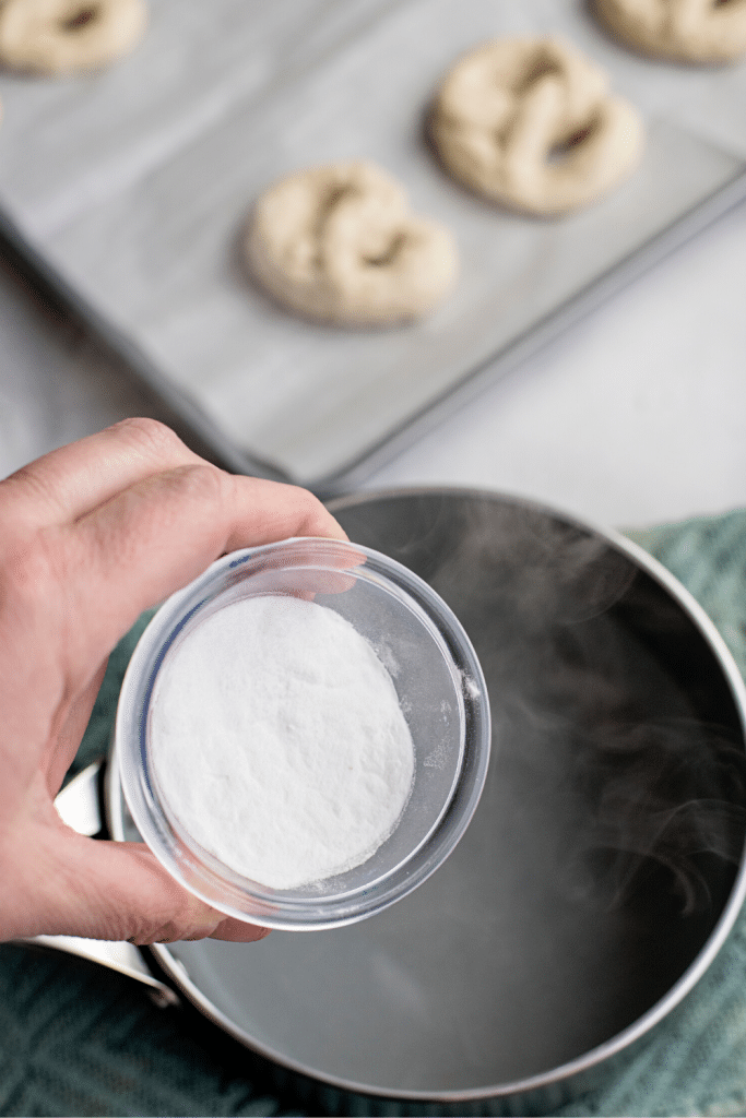 Soft Pretzels with Biscuit Dough 17 A hand holding a glass bowl with baking soda that's hovering over a pot of boiling water.