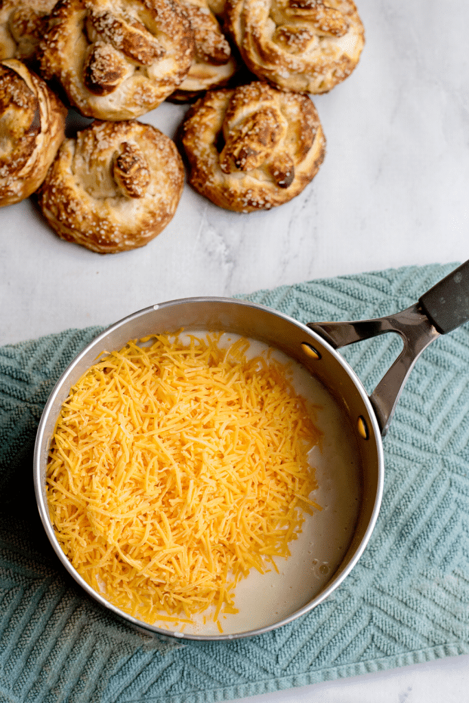Soft Pretzels with Biscuit Dough 23 Shredded cheese added to the pot, beside a batch of baked pretzels in the background.