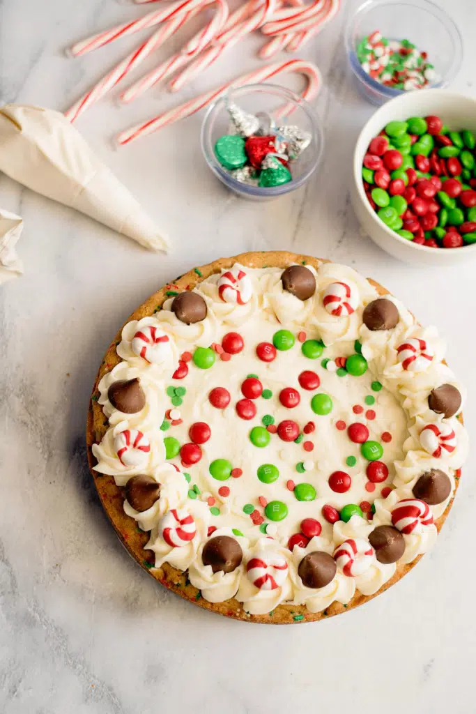 Overhead view of Christmas cookie cake topped with vanilla frosting and garnished with candy, piping bag with frosting, candy canes, and small bowls with Christmas candies on a white marble countertop.