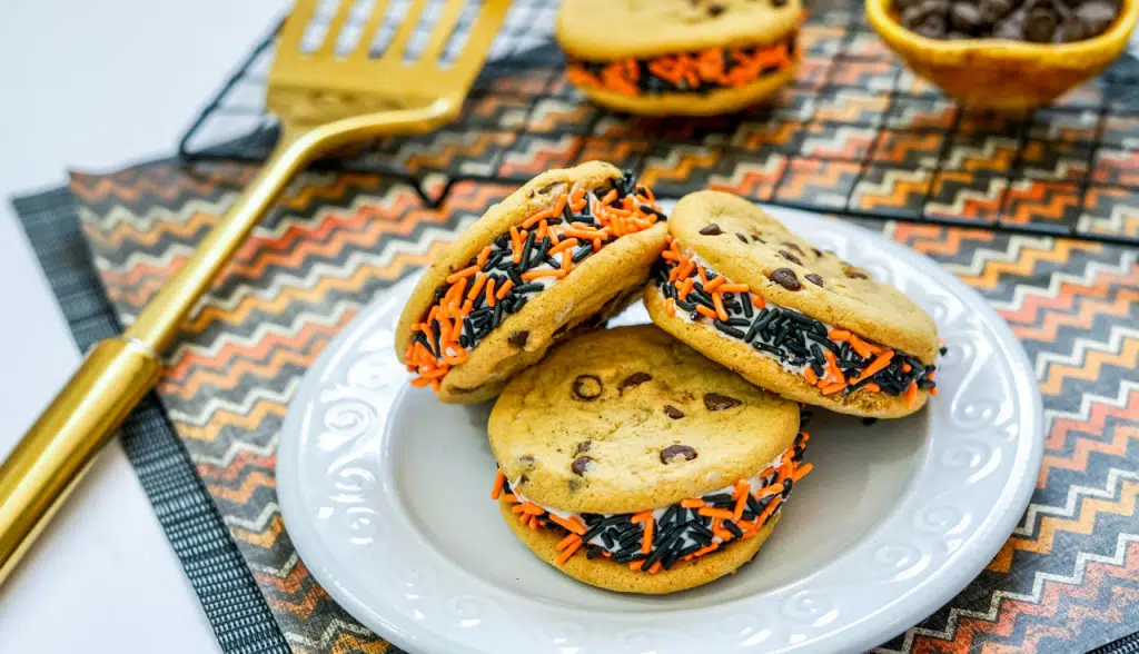 Three chocolate chip Halloween cookies on a white plate, additional decorative Halloween cookies on a cooling rack on a decorative mat.