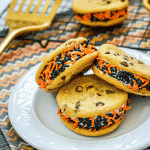 Three chocolate chip Halloween cookies on a white plate, additional decorative Halloween cookies on a cooling rack on a decorative mat.