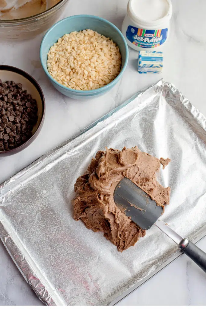 Chocolate marshmallow batter spread on a prepared baking sheet with a rubber spatula.
