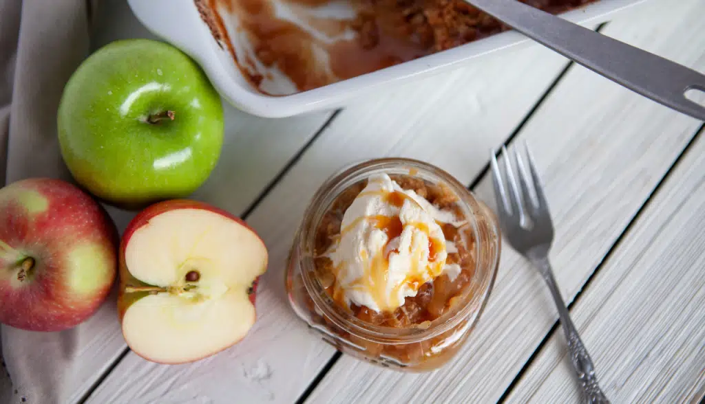 Overhead view of baking dish with easy apple crisp, glass jar with single serving of easy apple crisp, fresh apples, on a white wooden surface.