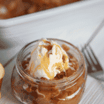 Easy apple crisp single serving in a glass jar with a dollop of whipped cream or scoop of vanilla ice cream, white baking dish with remaining easy apple crisp in the background, on a wooden countertop.