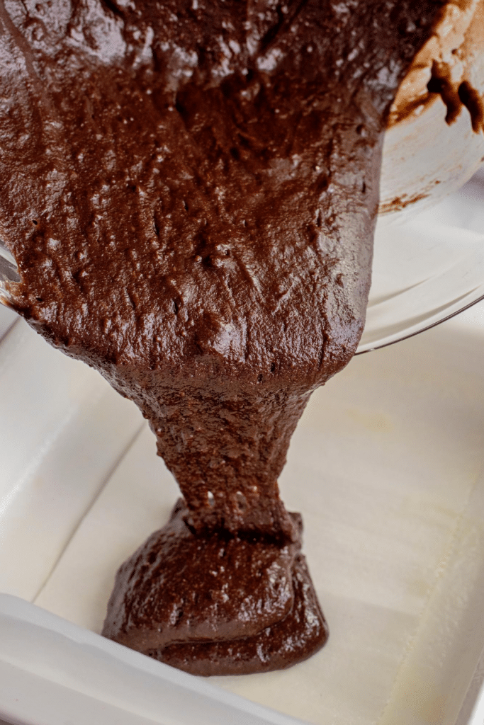 Thick chocolate brownie batter being poured from a glass bowl into a parchment-lined baking pan.