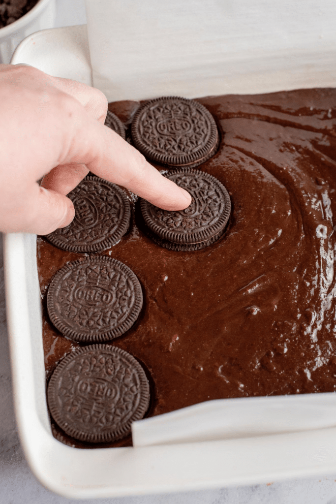 A hand presses Oreo cookies into chocolate brownie batter in a parchment-lined baking dish.