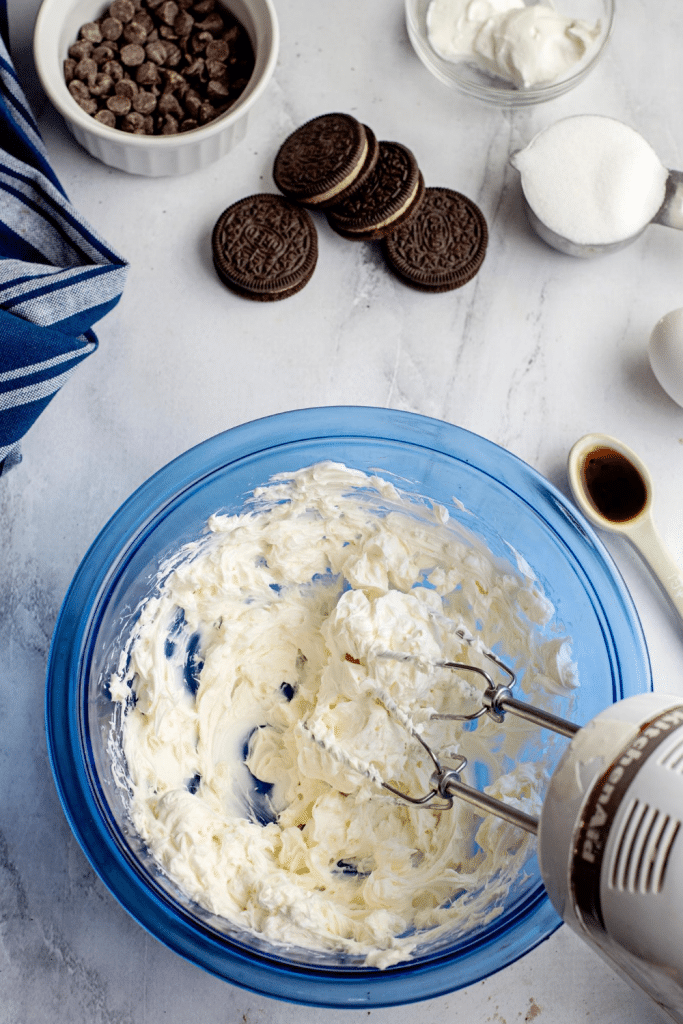 A hand mixer beats creamy mixture in a blue bowl surrounded by chocolate sandwich cookies, chocolate chips, a bowl of sugar, a spoon of vanilla, and a striped towel on a white countertop.