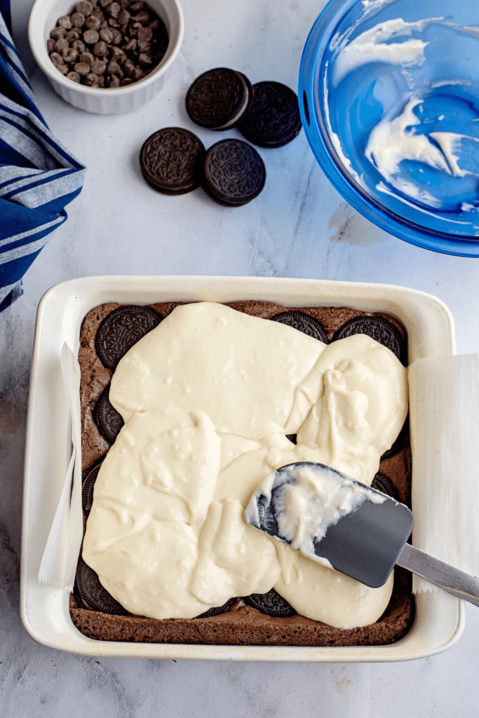 A baking dish with cookies at the bottom is being topped with creamy batter spread by a spatula. Nearby are a blue bowl with leftover batter, whole cookies, and a small dish of chocolate chips.