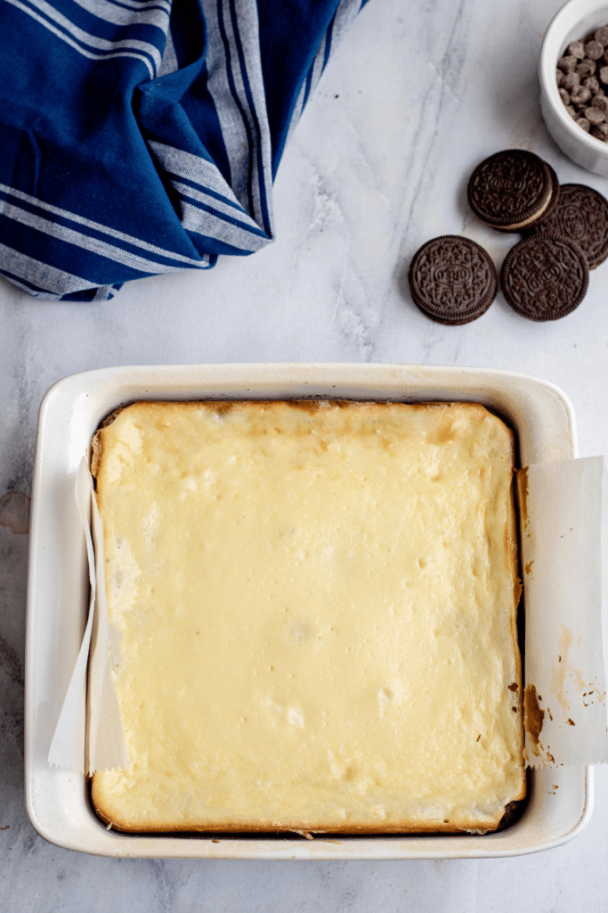 A square cheesecake in a white baking dish lined with parchment paper, next to three chocolate sandwich cookies, a bowl of chocolate chips, and a blue striped kitchen towel on a marble surface.