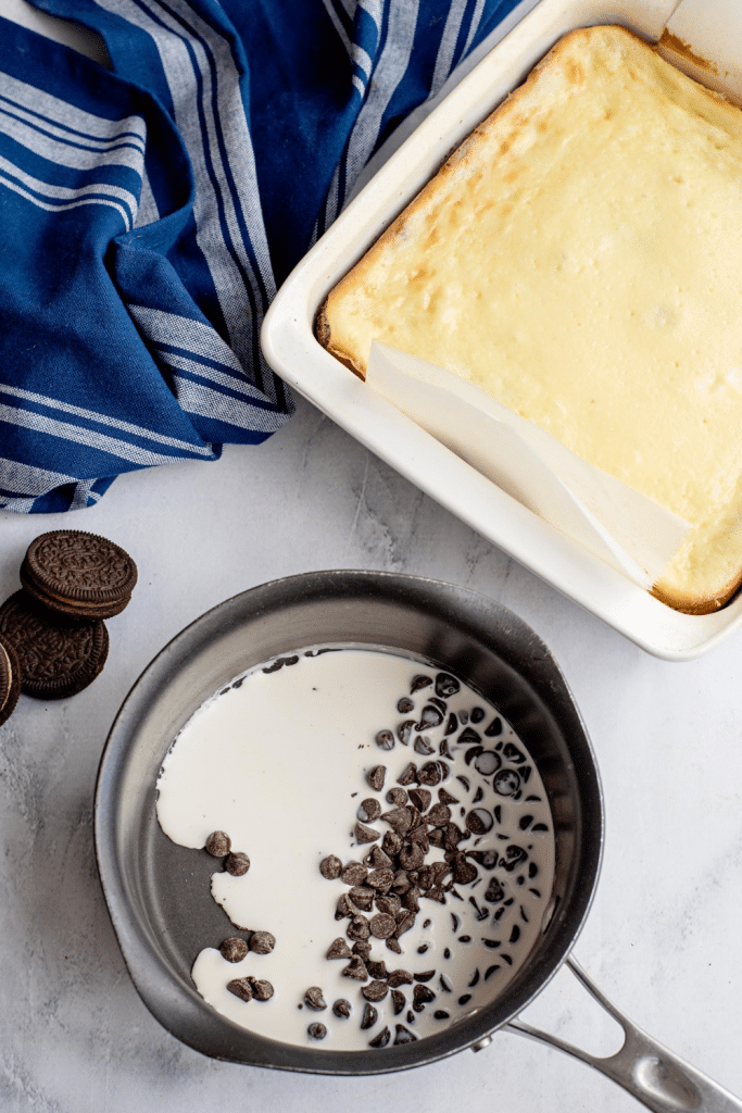 A saucepan with cream and chocolate chips sits next to a baked cheesecake in a white dish. A blue striped towel and two chocolate sandwich cookies are nearby on a light surface.