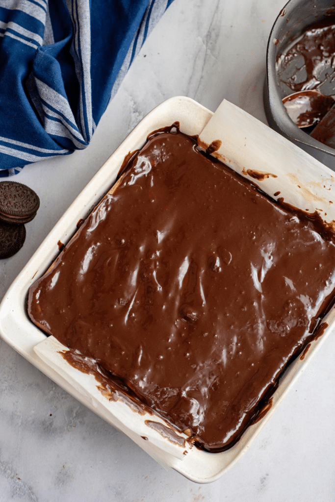 A square baking pan lined with parchment paper is filled with a layer of glossy chocolate batter. A few sandwich cookies and a blue striped cloth are nearby on a marble countertop.