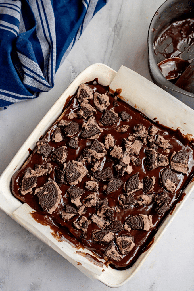 A square baking dish filled with chocolate brownies topped with pieces of chocolate sandwich cookies. A blue striped cloth and a saucepan with chocolate are next to the dish on a light countertop.