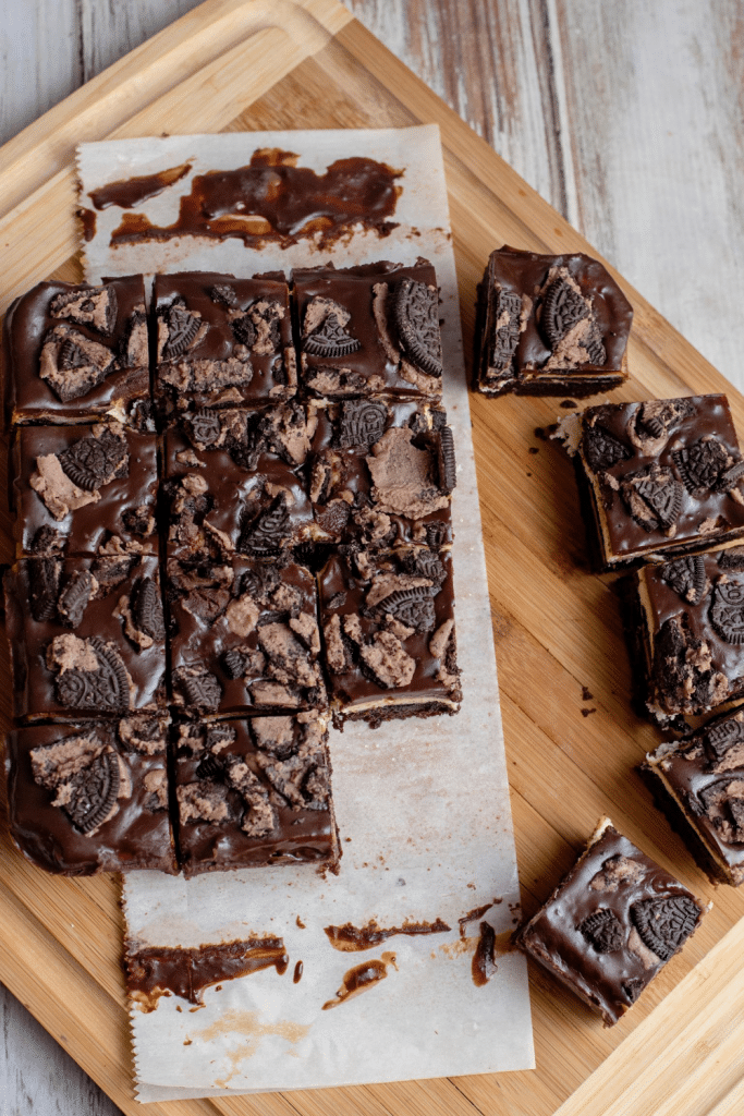 Squares of chocolate fudge topped with pieces of cookies are arranged on parchment paper atop a wooden cutting board, with several pieces separated from the main batch.