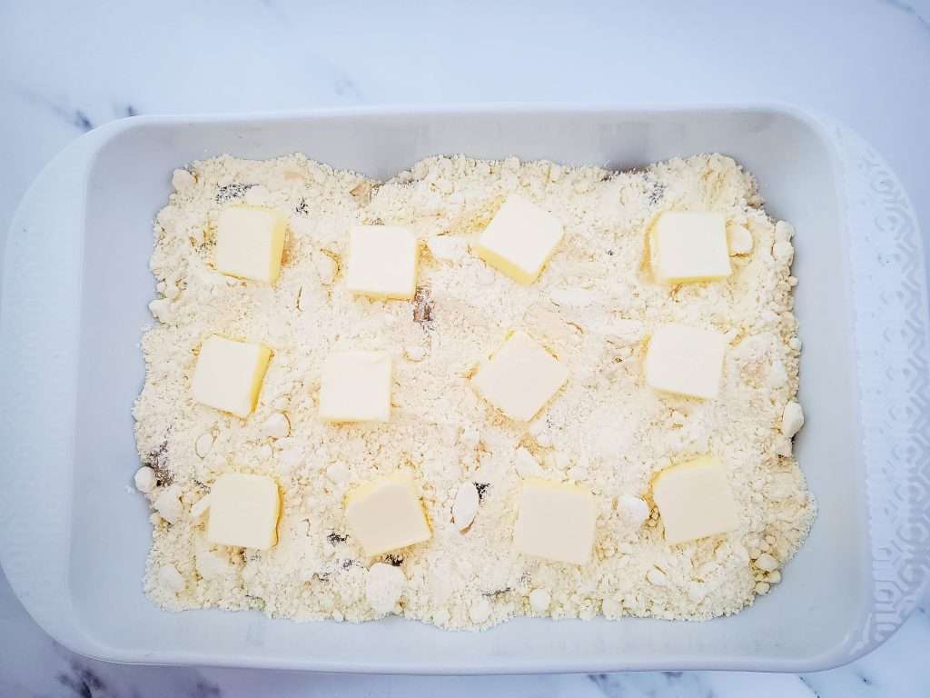 Apple Dump Cake 12 Sliced pieces of butter placed on the top of layered apple pie filling and cake mix in a white baking dish.