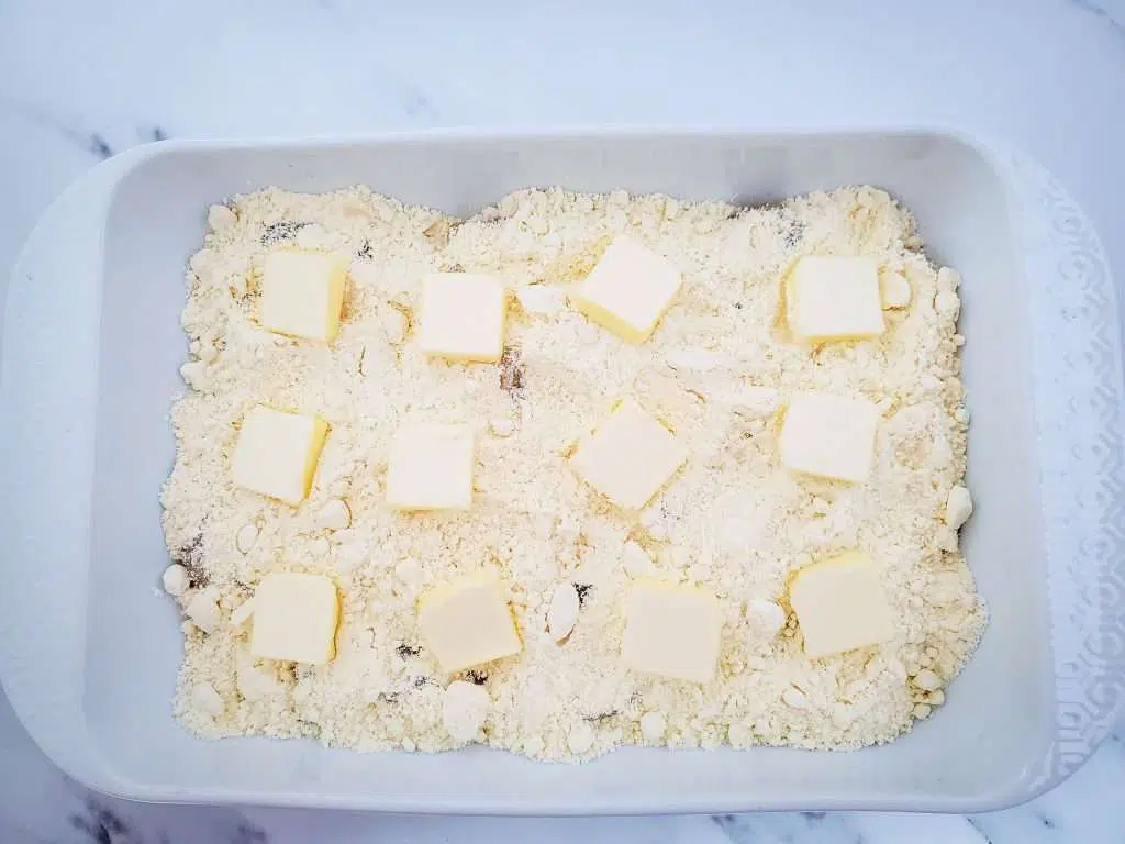 Sliced pieces of butter placed on the top of layered apple pie filling and cake mix in a white baking dish.