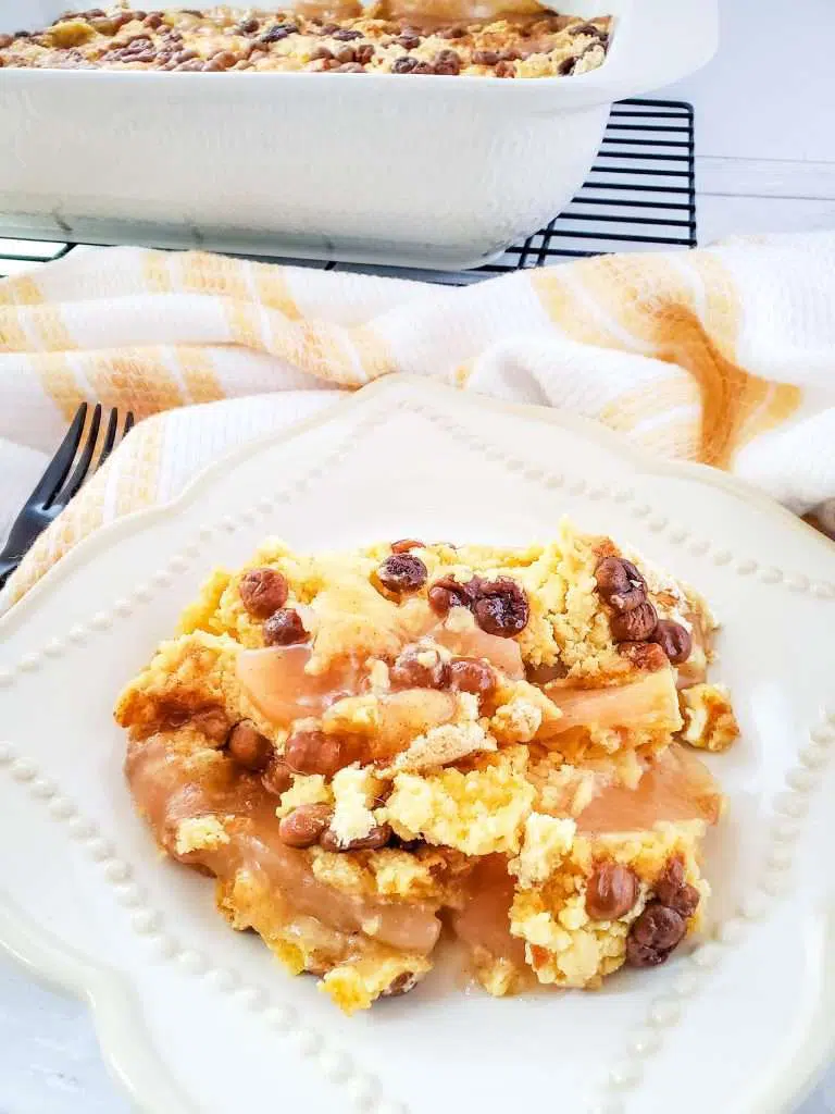 Apple dump cake on a serving plate, remaining apple dump cake in a baking dish on a cooling rack in the background, checkered linen cloth. 