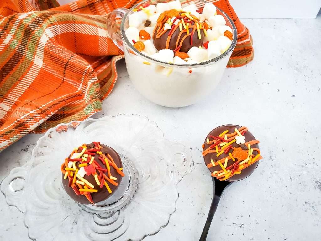 Fall-Themed Hot Chocolate Cocoa Bombs 2 fall-themed hot chocolate cocoa bombs sitting on a dish and in a mug