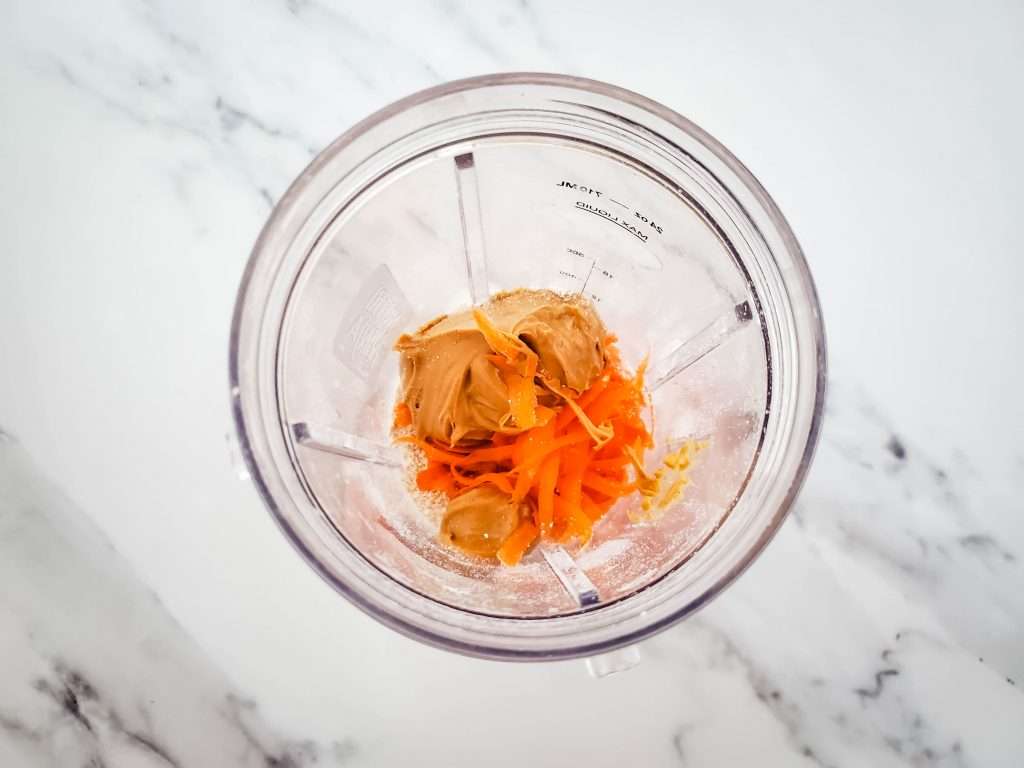 Blender container with peanut butter, ground oats, and shredded carrots.