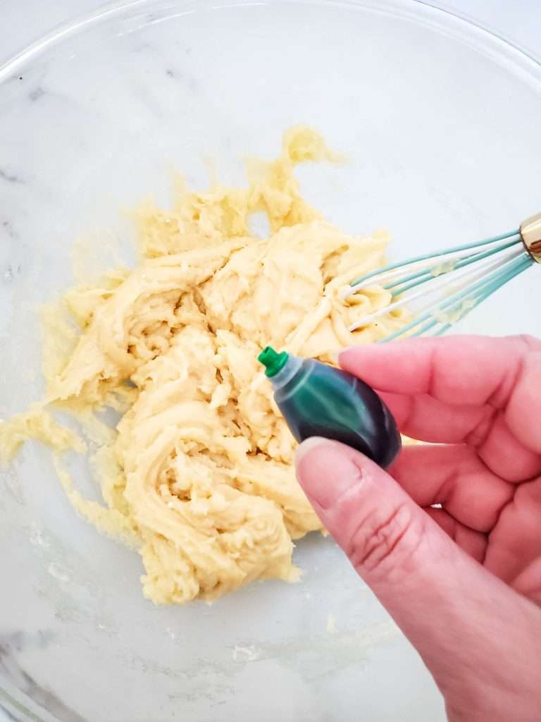Cake mix cookie dough in a bowl with a whisk and green food coloring being added.