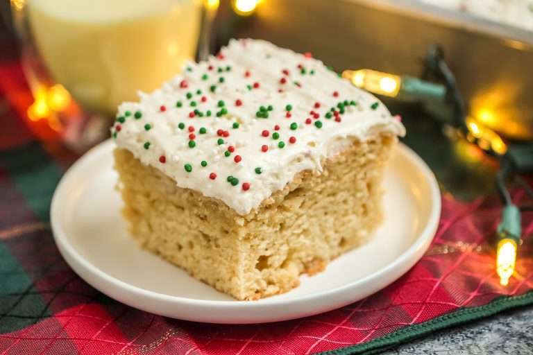 Close up of a slice of eggnog sheet cake on a white late with a glass of eggnog in the background.