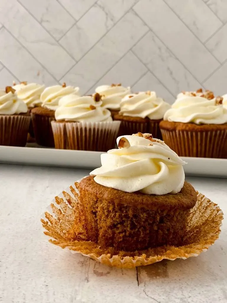 Close up of an apple cider cupcake unwrapped from the paper liner, plate of apple cider cupcakes in the background. 