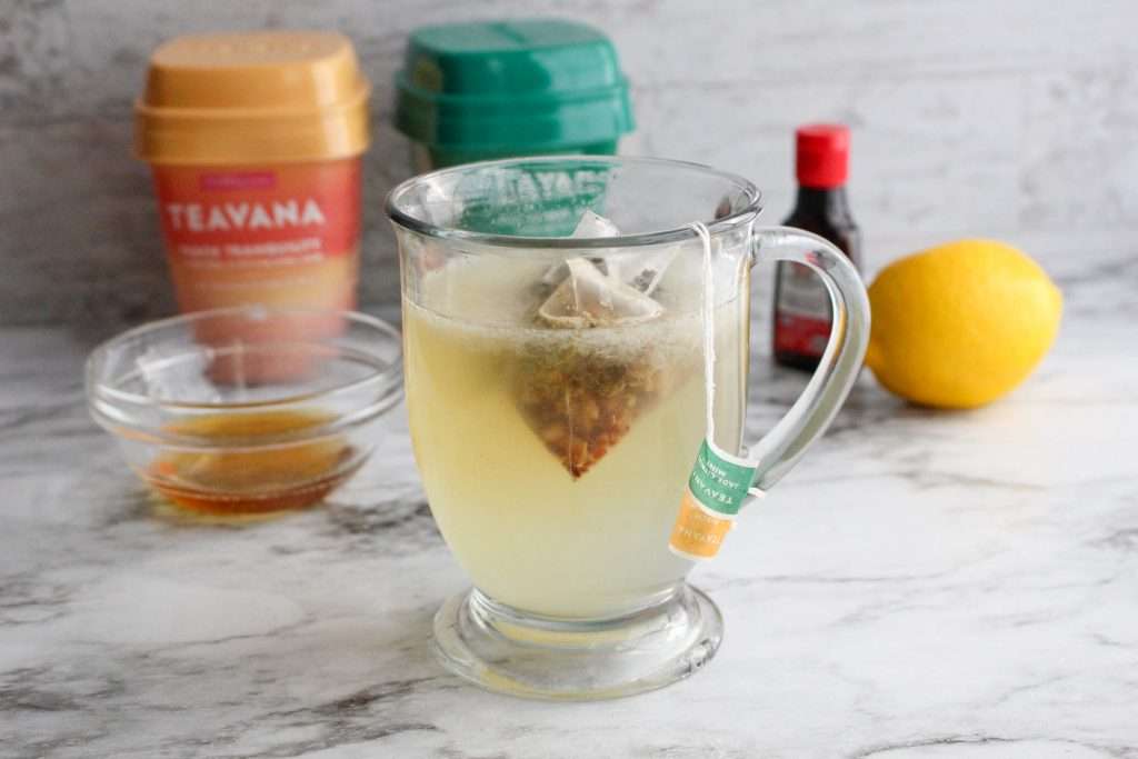 Starbucks Medicine Ball Tea Recipe (Copycat Recipe) 27 Glass with hot water, lemonade, and tea bags, copycat Starbucks Medicine Ball recipe ingredients in the background.