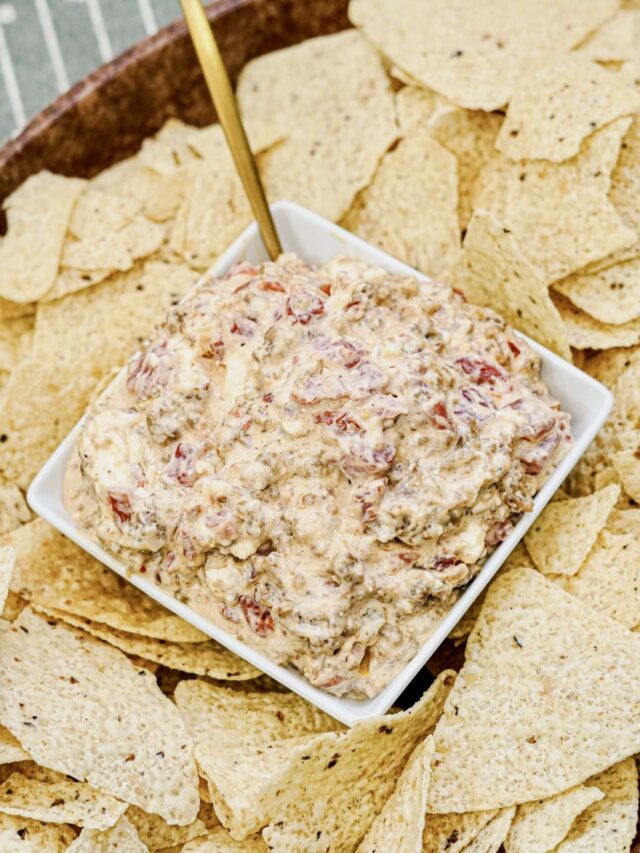 A square bowl of creamy dip with visible chunks of tomato and ground meat, surrounded by tortilla chips, with a gold spoon in the bowl.