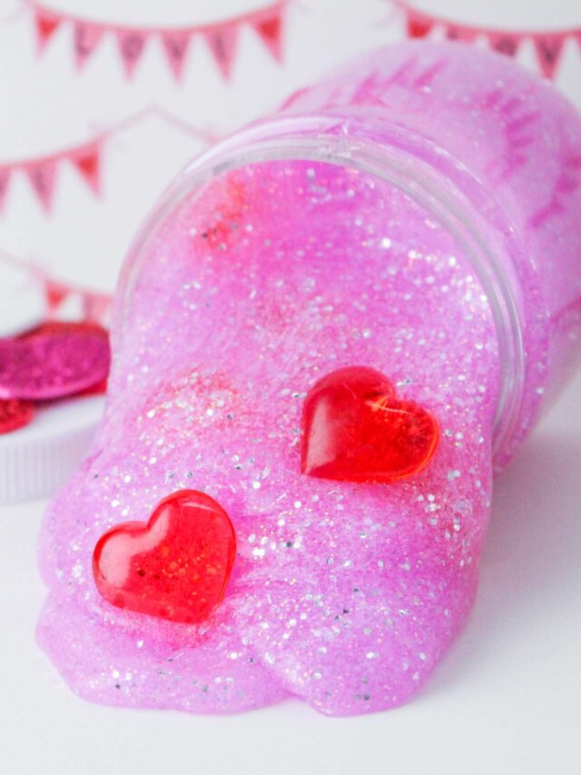 A clear jar of pink glitter slime spills out, with two red heart-shaped gems on top; a festive banner is blurred in the background.