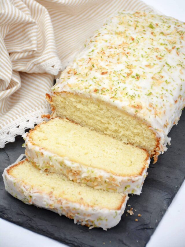 A loaf of glazed cake topped with shredded coconut and lime zest, partially sliced, sits on a dark slate surface with a striped cloth in the background.