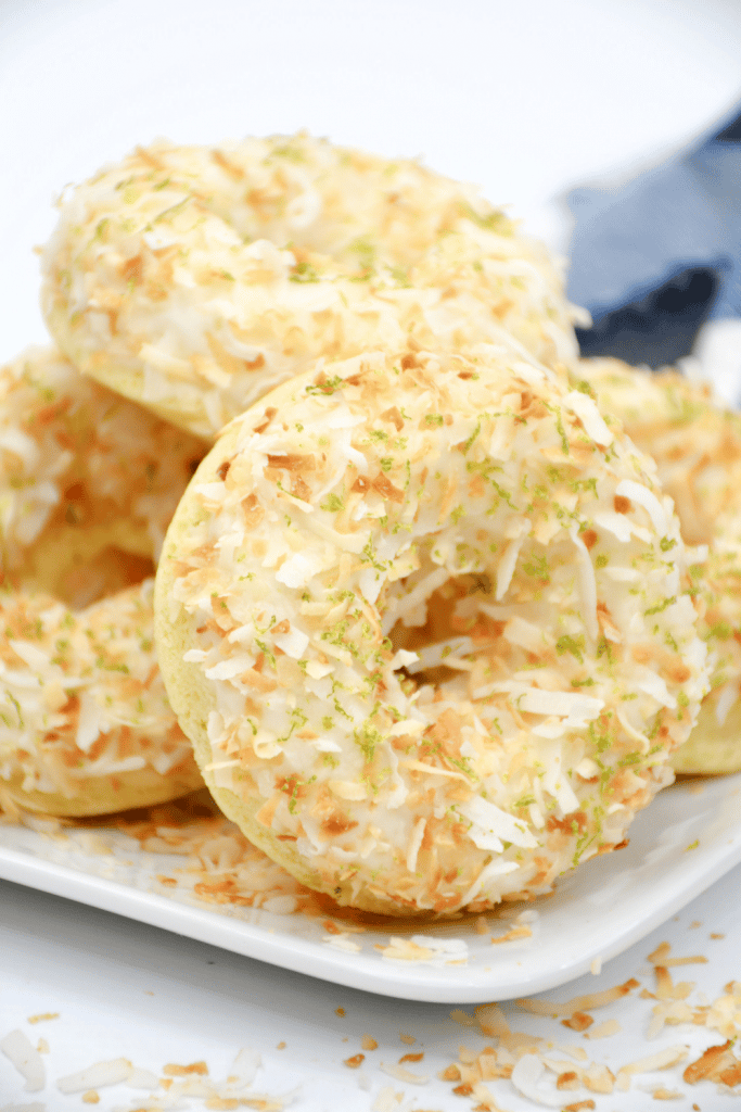 Baked Key Lime Coconut Donuts with Coconut Rum Glaze 4 A close-up of several baked donuts topped with shredded coconut and lime zest, stacked on a white plate with a blue napkin in the background.