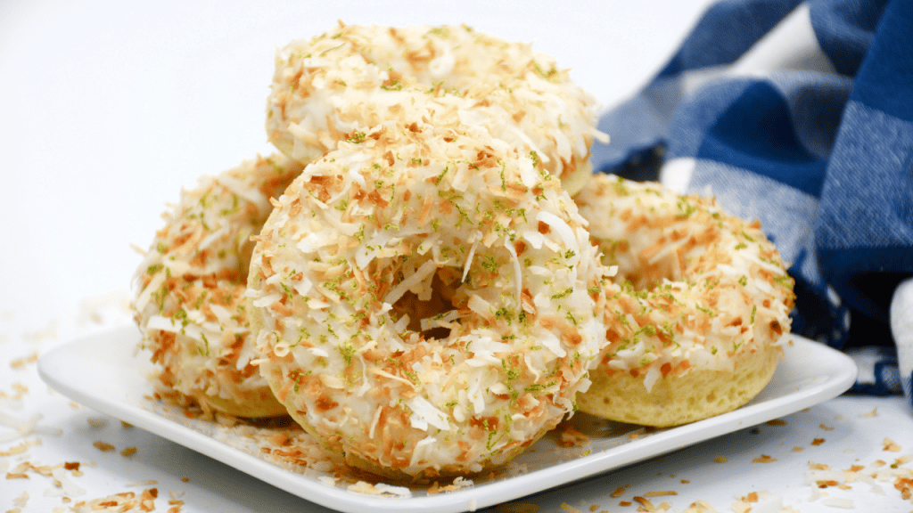 Baked Key Lime Coconut Donuts with Coconut Rum Glaze 3 A plate of baked donuts topped with white icing, toasted coconut flakes, and lime zest, with a blue and white checkered cloth in the background.