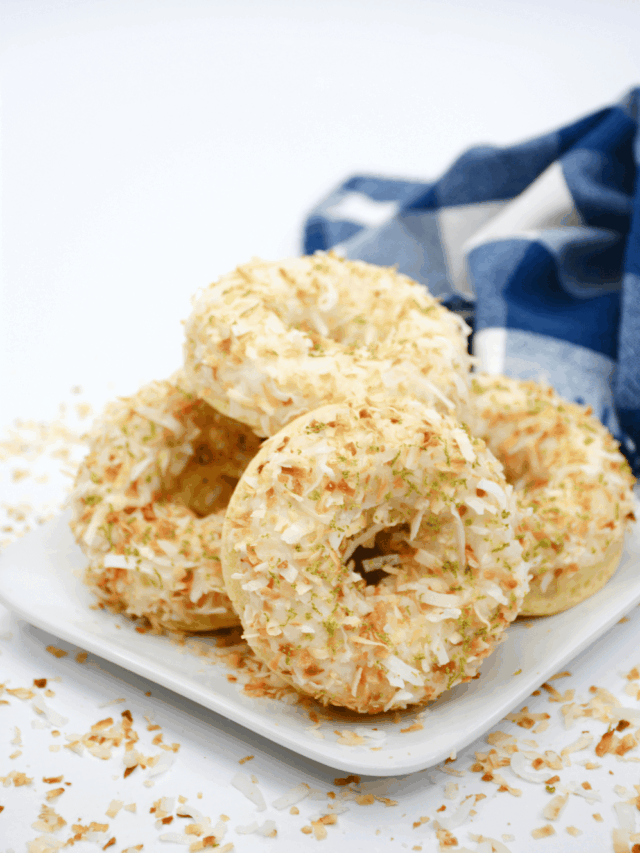 A stack of toasted coconut-covered donuts arranged on a white plate, with a blue and white cloth in the background.
