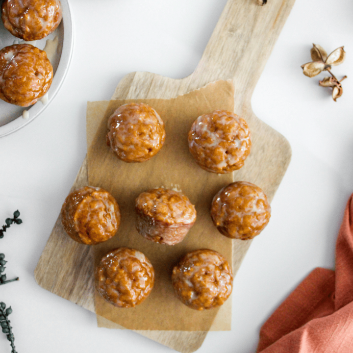 Overhead view of glazed pumpkin donut holes on a wooden kitchen board, jug of milk, additional donuts holes on a tray on a white countertop.